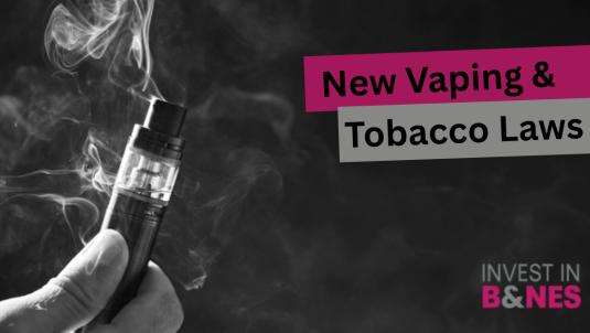 A close-up black-and-white image of a hand holding a vaping device, with vapour rising against a dark background. Text on the right reads "New Vaping & Tobacco Laws" in bold black lettering on pink and grey banners. The Invest in B&NES logo appears in the bottom-right corner.